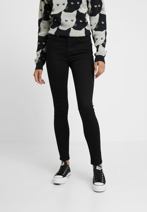 Person wearing black skinny pants, black and white cat-patterned sweater, and black low-top sneakers with white soles.