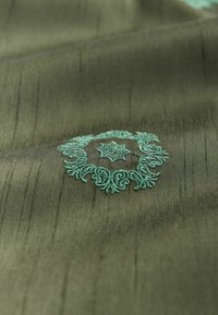 Olivgrüner Stoff mit glatter Textur, der in der Mitte ein kunstvolles türkisfarbenes Stickmuster in Form eines kreisförmigen Blumenmusters aufweist.