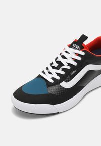 Baskets noires et bleues avec une tige texturée, des lacets blancs, un intérieur rouge contrastant et une semelle en caoutchouc blanche. Présente le logo Vans et des éléments de design uniques.