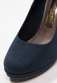 Zapato de tacón alto de ante azul marino con punta redonda y diseño de plataforma. Textura suave con un ligero brillo y un acento de logotipo dorado en el interior.