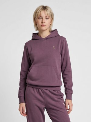 Lyshåret person stående frontalt iført en lilla hættetrøje og matchende bukser med lille logo på bryst og lår, neutral baggrund.