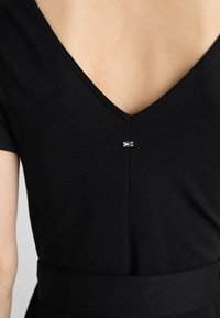 Widok z tyłu osoby noszącej czarną bluzkę z dekoltem w serek, z małą prostokątną przypinką w kolorach czerwonym, białym i niebieskim umieszczoną w górnym środku pleców.