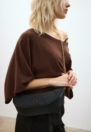 Blondinka v široki rjavi majici brez ramen in črnih hlačah z čipkastim robom, ki drži črno torbico z zlatim verižnim trakom.