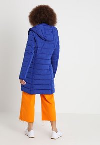 Persona de espaldas que lleva puesta una chaqueta acolchada con capucha azul, pantalones cortos naranjas brillantes y zapatillas blancas sobre un fondo llano.