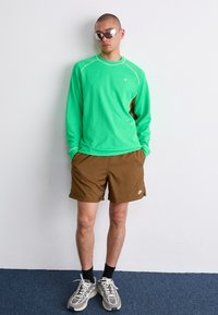 Grön långärmad skjorta med kontrasterande sömmar, bruna shorts och grå sneakers. Reflekterande solglasögon fulländar den sportiga looken mot en neutral bakgrund.