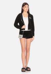 Felpa nera con zip, top senza maniche bianco, pantaloni corti in velluto nero, borsa argento con "Juicy" e sandali neri a ciabatta. Texture morbide ovunque.