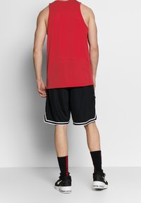 Débardeur rouge sans manches, short de basket-ball noir avec des rayures blanches, chaussettes noires avec des accents rouges, et chaussures de sport noires.