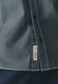 Maglia grigia con trama testurizzata e motivo a diamante dettagliato, cuciture bianche, bottoni neri e un'etichetta di tessuto con la scritta "DURABLE GOODS Lot. 182."