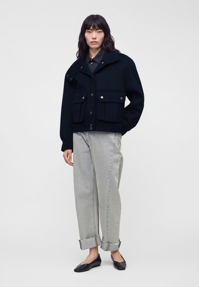 Femme portant une veste noire boutonnée avec de grandes poches, un jean gris clair avec revers, et des ballerines noires, debout devant un fond uni.