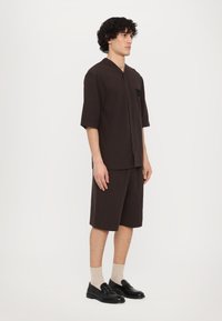 Men's dark brown short-sleeve shirt with a pocket, paired with matching shorts. Black loafers and beige socks complete the outfit.