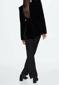 Czarna aksamitna marynarka z wyraźnie zarysowanymi ramionami, połączona z dopasowanymi czarnymi spodniami i butami na słupku. Gładka tekstura i jednolity kolor.