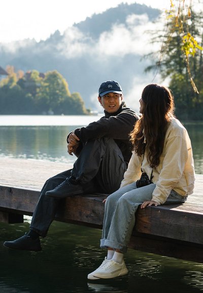 Dos personas están sentadas en un muelle de madera junto a un lago tranquilo. Una lleva una chaqueta negra y una gorra; la otra viste una chaqueta crema y jeans claros, con zapatillas blancas.