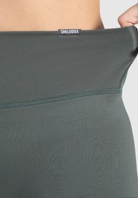 Cintura de ropa deportiva verde oscuro con una textura suave, que presenta una etiqueta negra con el texto "SMILODOX". La costura es visible a lo largo del borde.