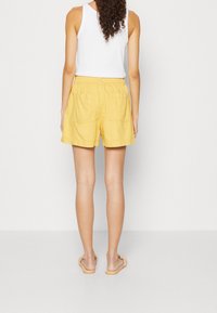 Kvinna med lockigt hår iförd en vit ärmlös topp, gula shorts med bakfickor och ljusbruna flätade sandaletter, stående vänd bort.