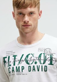 White cotton t-shirt featuring green printed text with aviation-themed graphics, round neckline, and short sleeves.