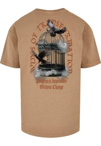 Beige bomulls t-skjorte med grafisk design av et fuglebur, to flygende fugler, og tekst som lyder "Vinger av transformasjon" i rødt.