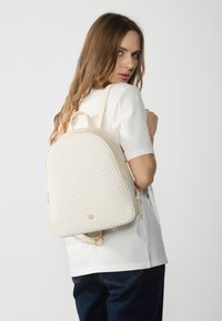 Fehér hátizsák texturált mintázattal és világos bézs díszítéssel. Arany színű fémalkatrészekkel és lekerekített tetejével rendelkezik, alkalmas hétköznapi használatra.