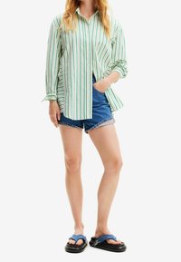 Persona che indossa una camicia a righe verdi e bianche con bottoni, pantaloni corti di jeans e sandali blu con suole spesse nere, in piedi con una mano in tasca.