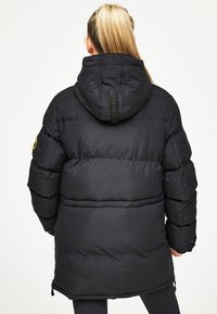 Melnā pūkām procesā žakete ar kapuci, ar izšūtām daļām, logotipa uzšuvumu uz kreisā piedurknes un gludu, spīdīgu apdari.