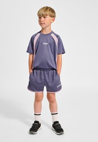 Lila sportset för barn, med en kortärmad tröja med rosa detaljer och matchande shorts, tillverkade i mjukt tyg. Svarta sneakers och randiga strumpor.