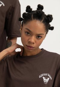 Young woman with bantu knot hairstyle wearing a brown t-shirt with white "Mystical Rising" butterfly logo on a white background.