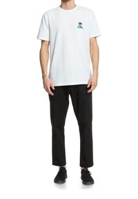 Witte katoenen T-shirt met een blauwe palmboomafbeelding op de borst, gecombineerd met zwarte katoenen broek en zwarte sneakers, staand in een neutrale houding.