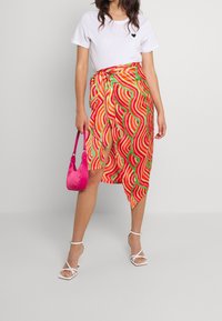 Jupe portefeuille colorée avec des motifs ondulés en rouge, orange et vert, associée à un t-shirt blanc uni et un sac à main rose. Talons transparents à brides.