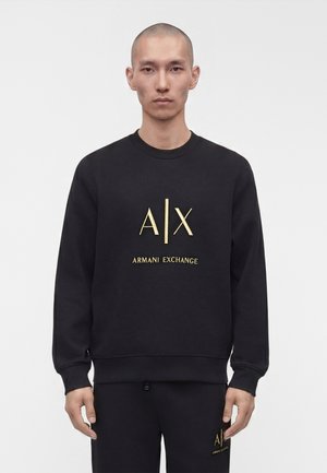 Uomo con testa rasata che indossa una felpa nera Armani Exchange e pantaloni abbinati con logo dorato, in piedi davanti a uno sfondo bianco uniforme.