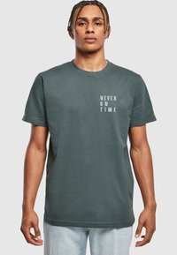 Mørkegrøn bomulds t-shirt med rund hals og korte ærmer, med "ALDRIG TIL TID" trykt i hvid på venstre brystområde.