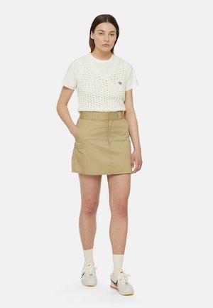 Camiseta blanca de manga corta superpuesta con un chaleco de punto, combinada con una falda beige. El conjunto se completa con unas zapatillas blancas y grises.