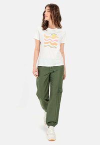 Witte t-shirt met kleurrijk golvend patroon; gecombineerd met olijfgroene cargobroek met zakken; afgemaakt met lichtgekleurde sneakers.