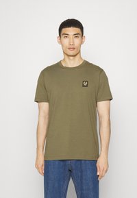 Tricou din bumbac de culoare verde măslin, cu guler rotund, mâneci scurte și o mică etichetă cu logo negru și aurie pe partea stângă a pieptului.
