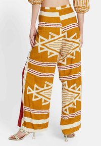 Pantalones de pierna ancha en mostaza con patrones geométricos en blanco y rojo. Hechos de una tela suave, con cintura alta y aberturas laterales.
