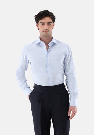 Uomo con capelli scuri che indossa una camicia azzurra infilata in pantaloni neri a vita alta, in piedi con una mano in tasca.