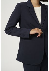 Osoba zapinająca ciemnoniebieską marynarkę na białej bluzce, nosząca cienką srebrną bransoletkę na lewym nadgarstku, na jednolitym jasnym tle.