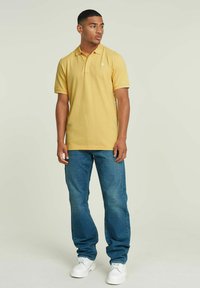 Maglietta polo gialla con colletto, maniche corte e dettagli del logo, abbinata a jeans blu e sneakers bianche. Tessuto a trama liscia.