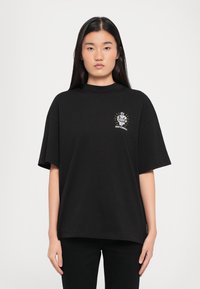 T-shirt nera oversize in cotone con collo alto e maniche corte, caratterizzata da un logo grafico bianco e oro sul petto.