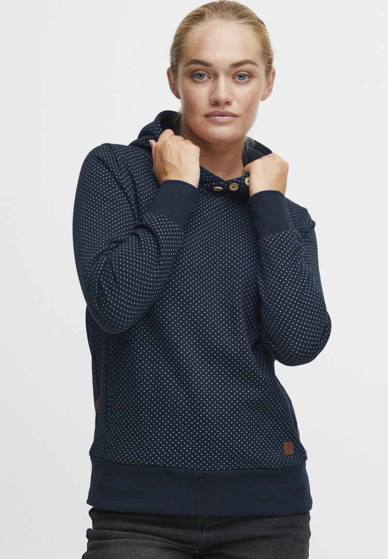 Sweatshirt à capuche bleu marine avec des pois blancs, en tissu texturé, présentant des boutons en bois au niveau du col et des poignets élastiques.