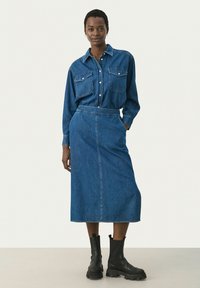 Jeans košulja s dva džepa na prsima i ovratnikom, u kombinaciji s dugom traper suknjom. Model nosi crne čizme do članaka. Svi komadi su u srednje plavoj nijansi.