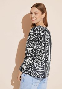 Blusa a fiori in bianco e nero con maniche a sbuffo, scollatura rotonda e pieghe sul retro; abbinata a jeans azzurri.