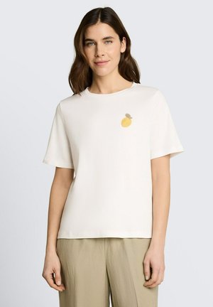 Femme aux cheveux bruns portant un t-shirt blanc uni à manches courtes avec une petite broderie de fleur jaune, associée à un pantalon beige clair.