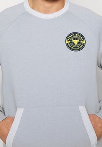 Sudadera gris claro hecha de una tela suave con acentos de rayas en blanco, que presenta un logotipo redondo en amarillo y azul en el pecho.