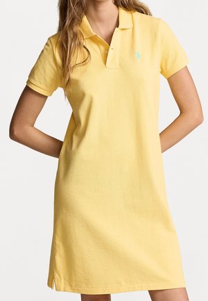 Femme portant une robe polo jaune à manches courtes, longueur genoux, avec un col et une patte de boutonnage, debout les bras derrière le dos.