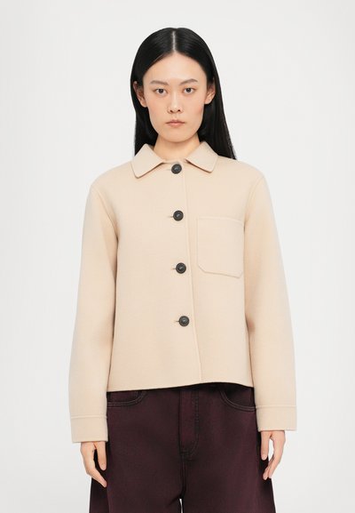 Veste en mélange de laine beige avec un col, une poche avant et des boutons noirs. Elle possède des manches longues et un design court. Portée sur un pantalon noir.