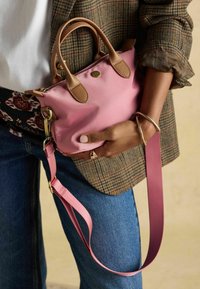 Malá ružová nylonová kabelka s hnedými koženými rukoväťami a detailmi, vybavená odnímateľným ružovým popruhom, držená na vzorovanom oblečení.