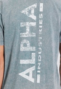 Hele sinine puuvillane t-särk, millel on julge, kulunud valge tekst, mis vertikaalselt vasakul küljel ütleb "ALPHA INDUSTRIES".