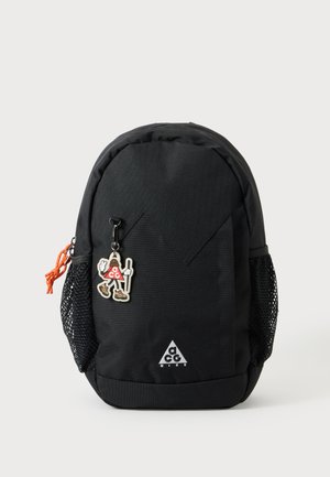 Sac à dos Nike ACG noir avec poches latérales en mesh, tirette de fermeture éclair orange et petit porte-clés personnage ACG accroché à l’avant.