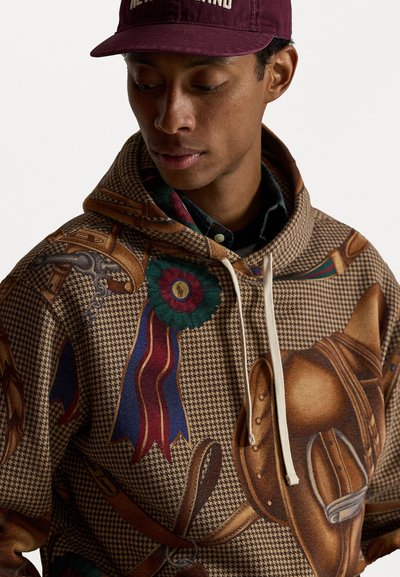 Jovem vestido com um hoodie castanho estampado com desenhos de equipamento equestre e um boné marrom com letras brancas.