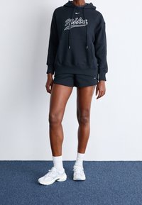 Svart hoodie med vit "Nike"-text, dragsko och känguruficka, kombinerad med svarta shorts och ljusa träningsskor.