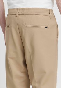 Beige trousers with a smooth texture, elastic waistband, and two back pockets. Features a small logo tag on the right pocket.